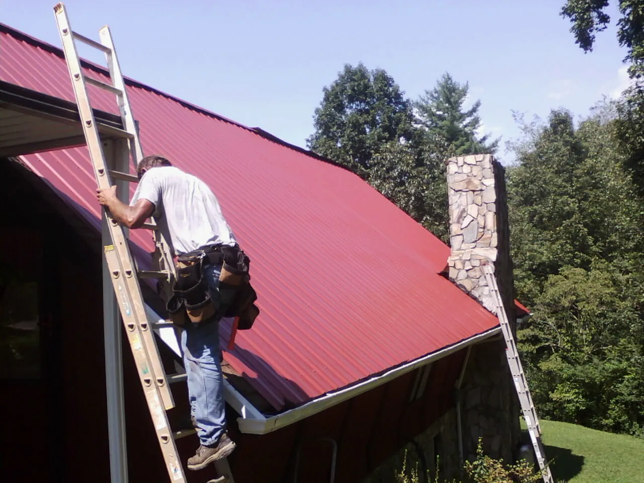 Roofer on a ladder beside a red metal roof and stone chimney — Soffit & Fascia Repair by Roofing Brothers in Maspeth